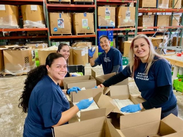 Four Marriott Vacations Worldwide associates in blue shirts and gloves smiling while working in a warehouse.
